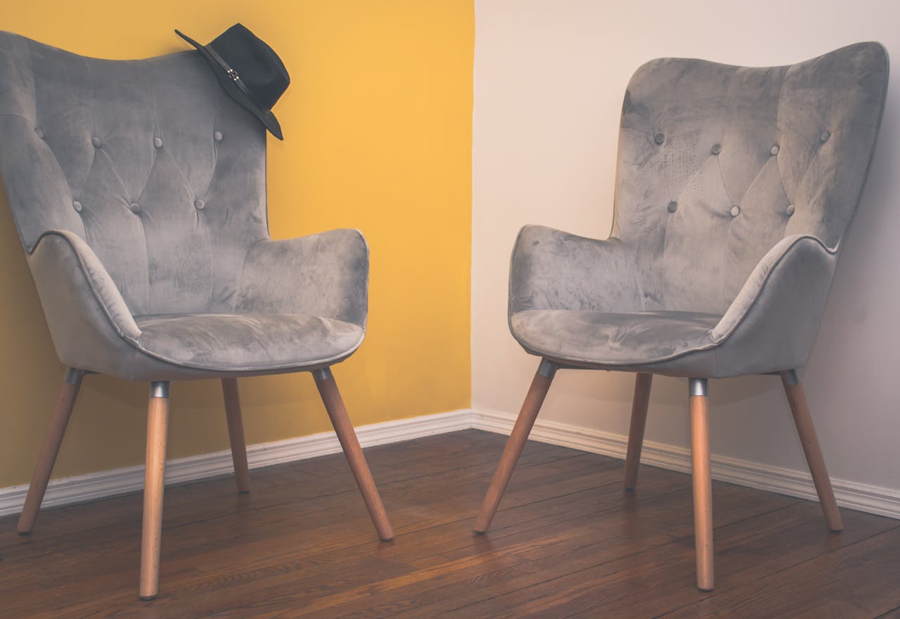 about-us Two contemporary gray armchairs in a minimalist interior with a yellow accent wall and wooden flooring.